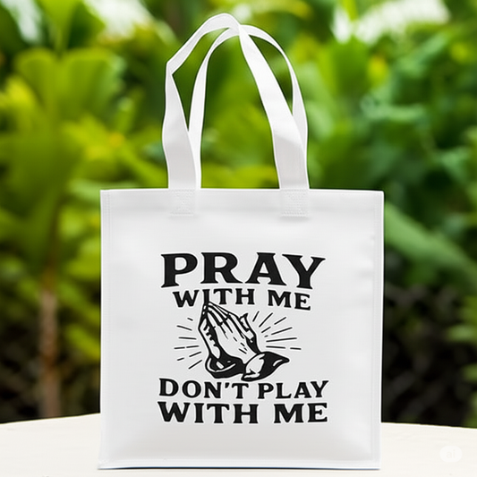 White tote bag with 'Pray with me, don't play with me' text and praying hands graphic on a green outdoor background.